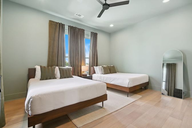Bright modern guest bedroom with two wooden platform beds, white linens and green-brown pillows, brown curtains, bedside lamp, ceiling fan, and arched floor mirror.