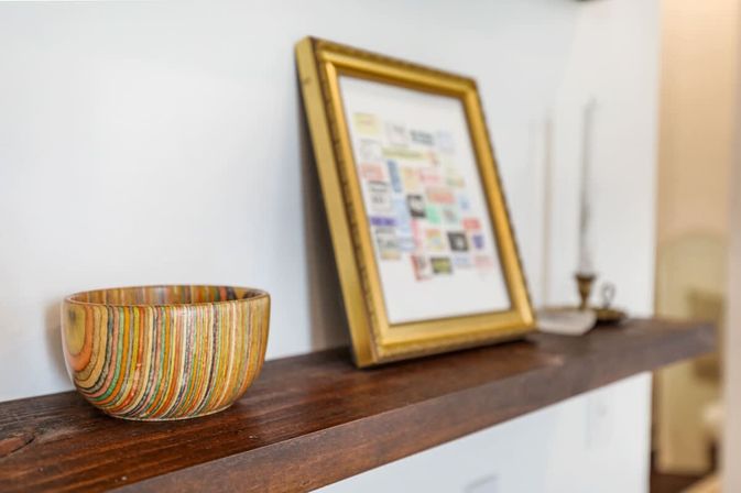 Cozy interior shelf with a colorful striped wooden bowl, a leaning gilded picture frame displaying a collage, and a vintage candlestick on a dark wood ledge.