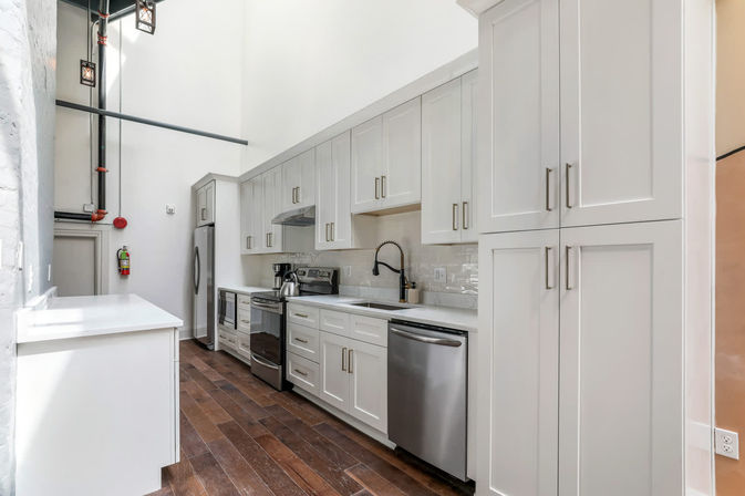 Bright urban loft galley kitchen with high ceilings, exposed industrial pipes and hanging lanterns, white shaker cabinets, quartz countertops, stainless steel appliances, gooseneck faucet, subway tile backsplash and dark hardwood floors.