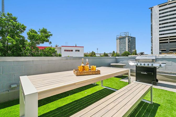Sunny urban rooftop patio with modern wood picnic table and benches on green turf, wicker tray with drinks, stainless steel gas grill and downtown buildings under a clear blue sky.