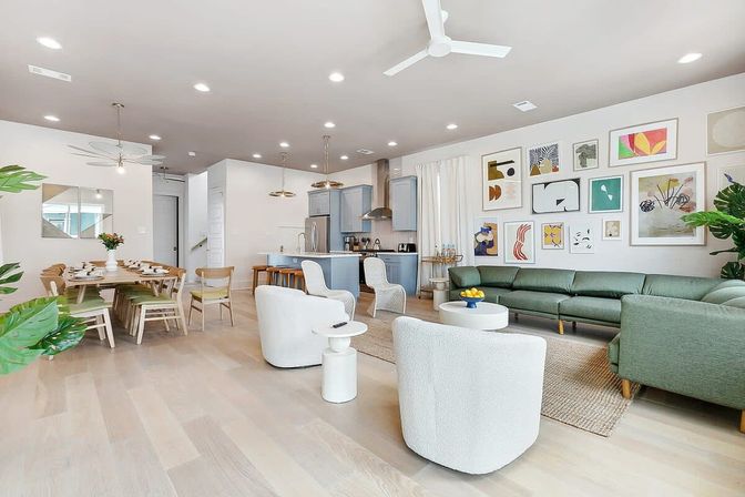 Bright, airy open-concept living room and kitchen with light wood floors, green sectional sofa, white swivel chairs, gallery wall, dining table and island with pendant lights.