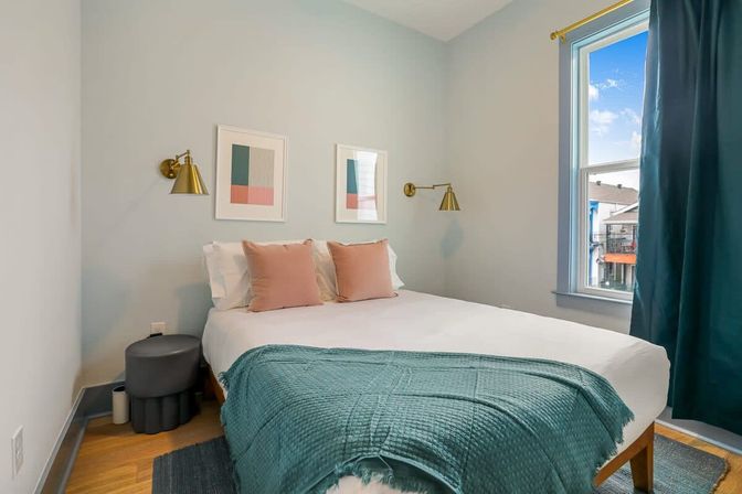 Cozy urban apartment bedroom with white bedding, teal throw, blush pillows, brass wall sconces, minimalist framed art and a window overlooking neighboring buildings.