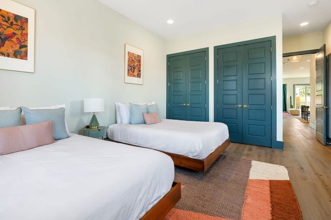 Coastal-inspired bright twin bedroom with white linens, pastel throw pillows, wood platform beds, teal double-paneled closet doors, bedside lamp and framed art, and a colorful woven rug on hardwood floors.