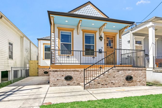 Cheerful raised cottage with white siding, mustard trim and turquoise porch ceiling, brick foundation, wrought-iron railing, wooden front door and concrete driveway on a sunny day.