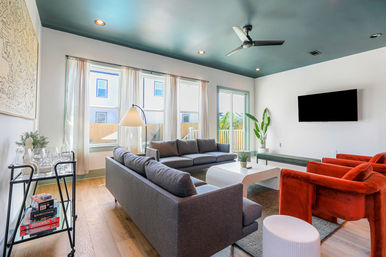 Sunlit modern living room with teal ceiling, gray sectional sofa, white coffee table, red velvet armchairs, wall-mounted TV, sheer curtains and hardwood floors.