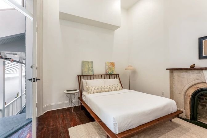 Bright minimalist bedroom with a low wooden platform bed dressed in white linens and patterned lumbar pillow, tall white walls, dark hardwood floors, small round side table and floor lamp, marble fireplace mantel, and an open door to an exterior balcony.
