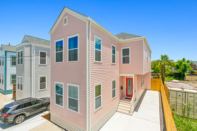 Two-story pink clapboard house with white-trim windows and red front door, concrete driveway with parked SUV, neighboring pastel homes and palm trees under a clear blue sky