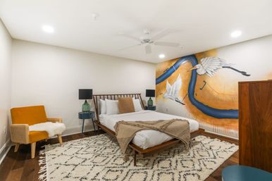 Modern bedroom with a wooden mid-century bed, textured beige throw and patterned rug, mustard accent chair and green glass bedside lamps, plus a large warm-toned wall mural of a winding blue river with two flying white herons.