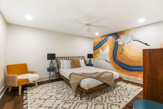 Modern bedroom with a wooden mid-century bed, textured beige throw and patterned rug, mustard accent chair and green glass bedside lamps, plus a large warm-toned wall mural of a winding blue river with two flying white herons.