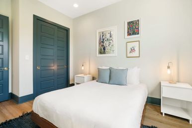 Modern minimalist bedroom interior with white bed and two light-blue pillows, white nightstands with exposed-bulb lamps, botanical wall art, navy-blue sliding closet doors and trim, and light wood floors — bright, cozy design.