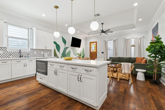 Bright open-plan kitchen and living room with a large white island, brass globe pendant lights, patterned backsplash, dark hardwood floors, green sofa, rattan chairs, leafy wall mural and potted fiddle-leaf fig.