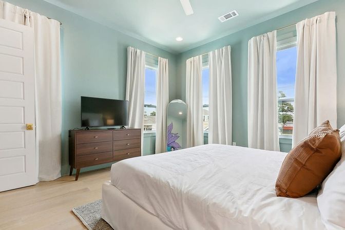 Sunlit airy bedroom with seafoam-green walls, white linens and tan accent pillow on a bed, three tall windows with white curtains, mid-century wooden dresser with flat-screen TV, arched full-length mirror and light hardwood floors.