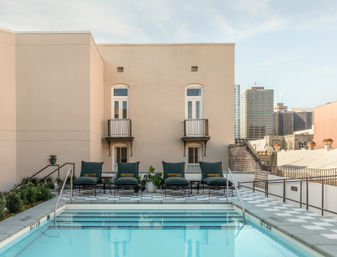 Sunlit rooftop pool oasis with turquoise water, checkered tile deck and green lounge chairs facing a pale stucco building with small balconies and a distant urban skyline.