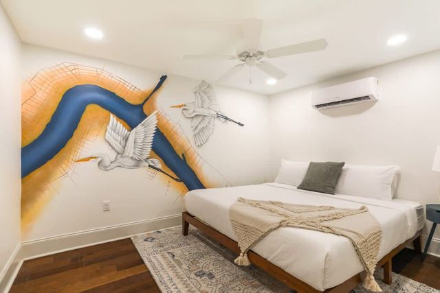 Bright modern bedroom with a coastal-inspired wall mural of two white herons flying over a stylized blue river and orange accents, hardwood floors, patterned rug, white bed with textured throw, ceiling fan and wall-mounted AC.