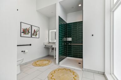 Bright modern accessible bathroom with walk-in shower lined in dark green subway tiles, stainless grab bars, wall-mounted sink, toilet, botanical framed prints and yellow round rugs