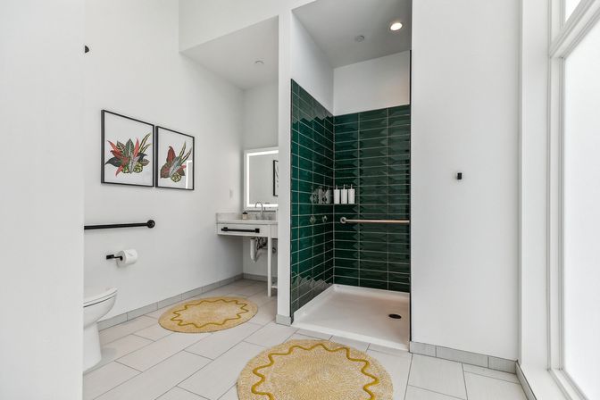 Bright modern accessible bathroom with walk-in shower lined in dark green subway tiles, stainless grab bars, wall-mounted sink, toilet, botanical framed prints and yellow round rugs