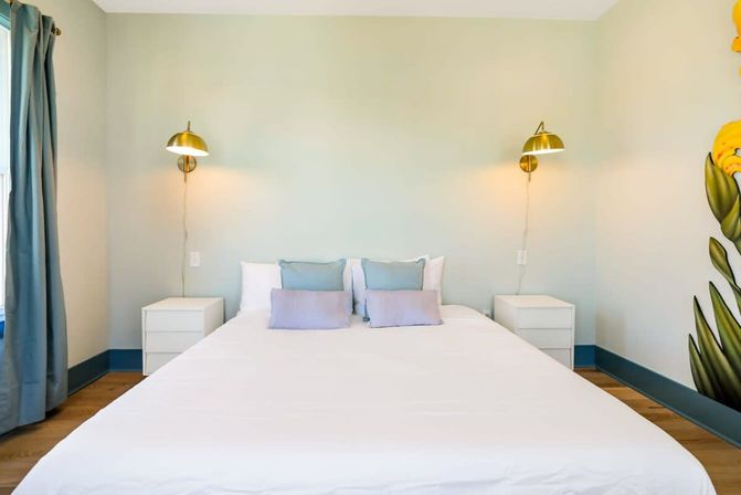 Bright minimalist bedroom interior with a white bed and linens, pastel blue and lavender throw pillows, brass wall sconces above matching white nightstands, blue curtains and a floral mural accent.