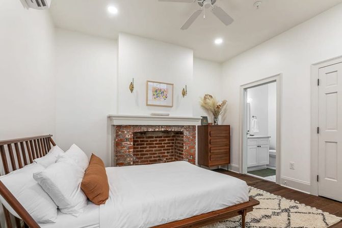 Bright minimalist bedroom with wooden slatted bed, white bedding and rust accent pillow, exposed red brick fireplace with white mantle, mid-century dresser with pampas grass, patterned rug, ceiling fan and doorway to an ensuite bathroom.