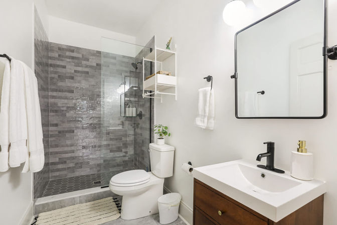 Bright modern bathroom with gray subway-style tile walk-in shower behind a glass door, white toilet, wooden vanity with white sink and black faucet, black-framed mirror, wall shelves and white towels.
