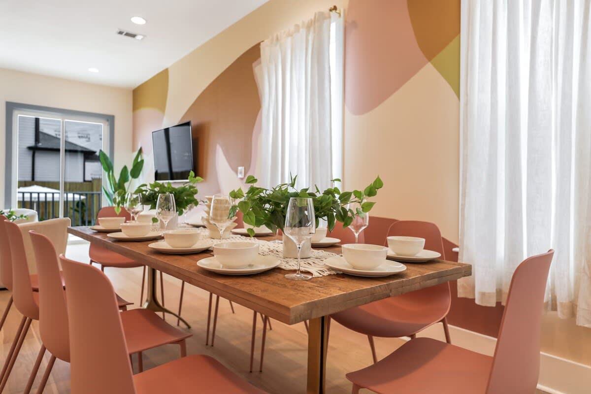Bright modern dining room with rustic wood table set for six, blush-pink chairs, white dinnerware and wine glasses, green centerplants, abstract peach wall mural, sheer curtains and sliding glass door letting in natural light.