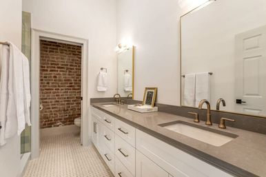 Bright modern bathroom with double undermount sinks, gray quartz countertop, brass faucets, white shaker cabinets, large mirror, patterned tile floor and a toilet alcove with exposed brick accent wall.