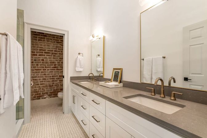 Bright modern bathroom with double undermount sinks, gray quartz countertop, brass faucets, white shaker cabinets, large mirror, patterned tile floor and a toilet alcove with exposed brick accent wall.