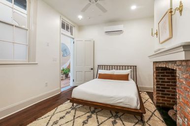 Cozy airy modern bedroom with wooden platform bed, white linens and brown lumbar pillow, exposed red brick fireplace, patterned area rug on dark hardwood floors, white walls, ceiling fan, and open doorway to a plant-filled adjacent room.