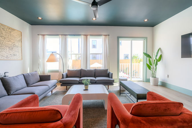 Sunlit modern living room with gray sofas, two coral-red velvet armchairs, white coffee table with plant, arched floor lamp, ceiling fan, and sliding glass door to a balcony.