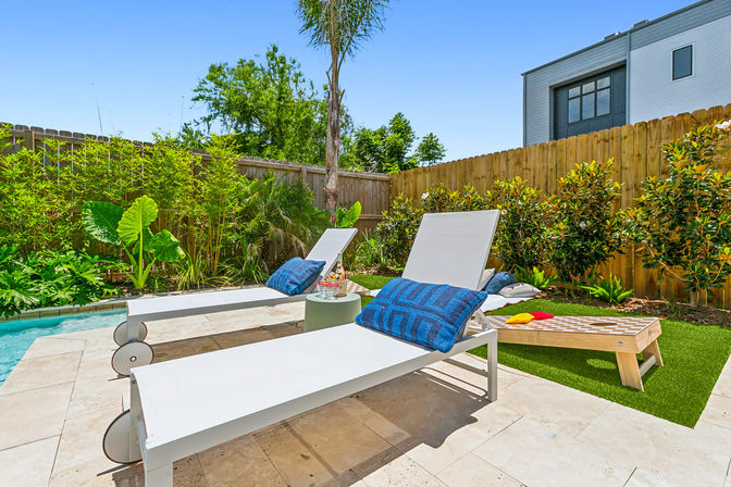 Sunny backyard poolside patio with two white lounge chairs topped with blue patterned pillows, small round side table with drinks, cornhole board on green turf, lush tropical plants and wooden fence