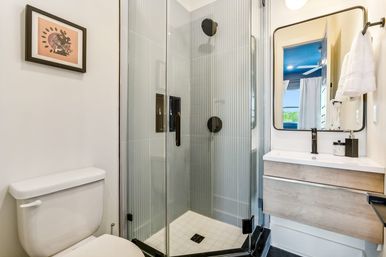 Modern compact residential bathroom with corner frameless glass shower featuring vertical ribbed gray tiles, white toilet, wood-tone floating vanity, black matte fixtures and rectangular mirror.