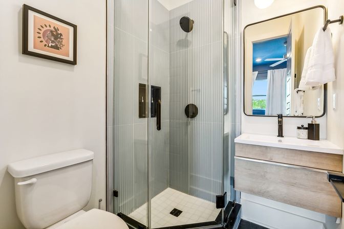 Modern compact residential bathroom with corner frameless glass shower featuring vertical ribbed gray tiles, white toilet, wood-tone floating vanity, black matte fixtures and rectangular mirror.