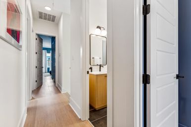Contemporary residential hallway interior with light wood floors, white paneled doors and walls, an open bathroom showing an oak vanity with black faucet and round mirror lights, and a blue-accented bedroom visible at the end of the hall.