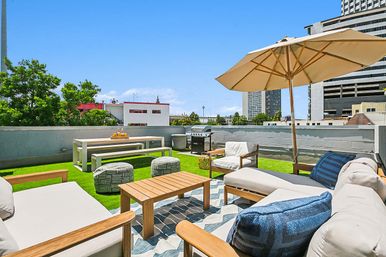 Sunny urban rooftop terrace with cushioned lounge seating, wooden coffee table, large beige umbrella, picnic bench, grill and green turf overlooking downtown buildings