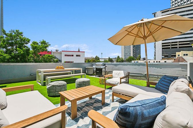 Sunny urban rooftop terrace with cushioned lounge seating, wooden coffee table, large beige umbrella, picnic bench, grill and green turf overlooking downtown buildings