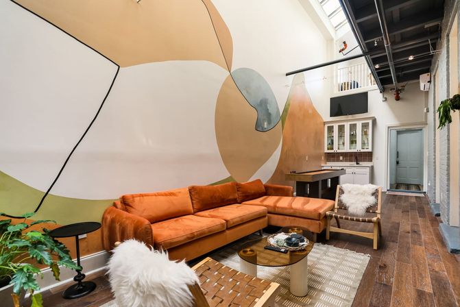 Bright modern loft living room with rust-orange velvet sectional, sweeping abstract wall mural, skylight, hardwood floors, woven chairs with sheepskin throws and a compact kitchenette