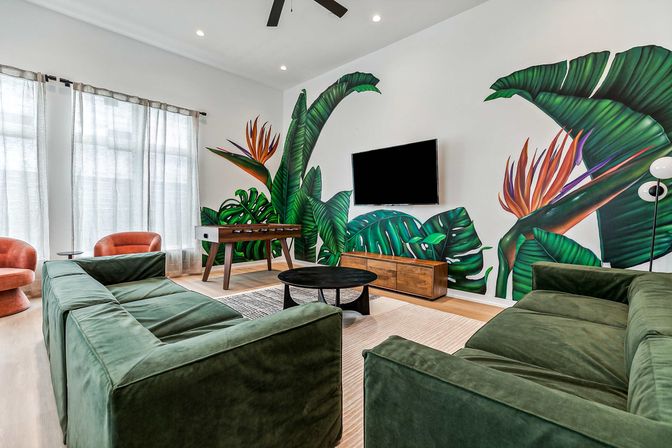 Sunlit contemporary living room with a giant tropical leaf mural, green velvet sofas, wall-mounted TV, wooden media console, foosball table and rust-colored accent chairs beneath sheer curtains.