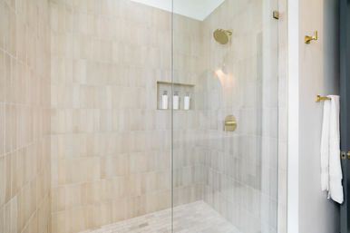 Bright modern glass shower with beige subway tiles, brass rain showerhead and controls, recessed niche holding three white dispensers, light-colored tile floor and a white towel on a wall hook