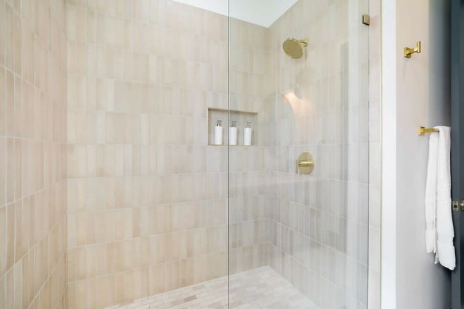 Bright modern glass shower with beige subway tiles, brass rain showerhead and controls, recessed niche holding three white dispensers, light-colored tile floor and a white towel on a wall hook