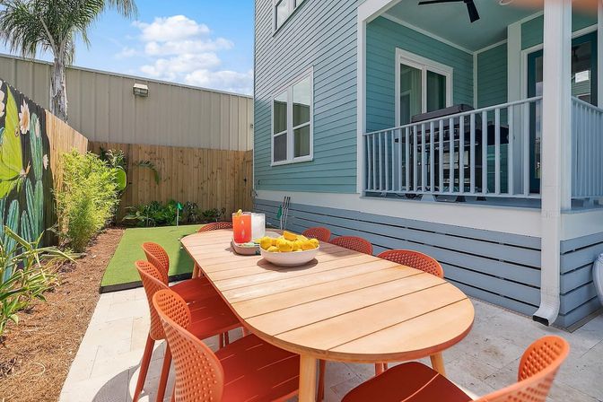 Sunny backyard patio with a wooden outdoor dining table surrounded by orange chairs, bowl of lemons and a pitcher, blue coastal-style house with covered porch, small artificial turf putting green and painted garden fence.