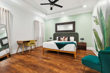 Modern bedroom interior with king bed featuring a dark green velvet headboard against a light green accent wall, warm wood floors, mid-century desk and chair, teal lounge chair, tropical leaf wall mural, ceiling fan and full-length mirror.