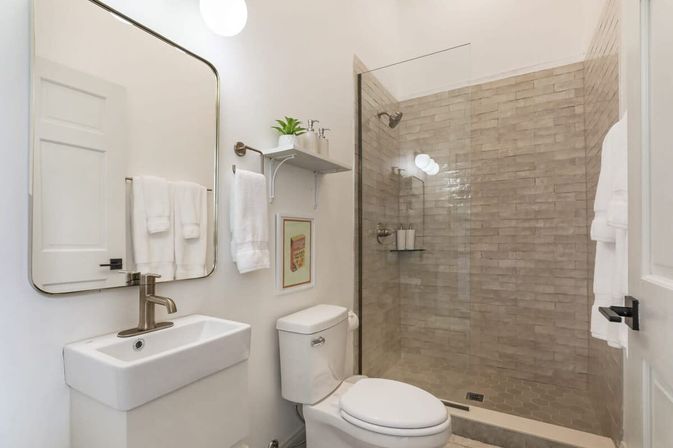 Bright modern small bathroom with a white sink and brushed nickel faucet under a rounded-rectangle mirror, toilet, glass-enclosed walk-in shower with warm beige subway tiles, white towels and a small shelf plant—spa-like and compact.