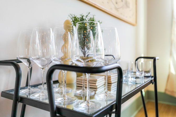 Sparkling wine glasses and tumblers neatly arranged on a black metal bar cart with mirrored shelf, stacked coasters, decorative candle and small potted plant in a bright modern dining area.
