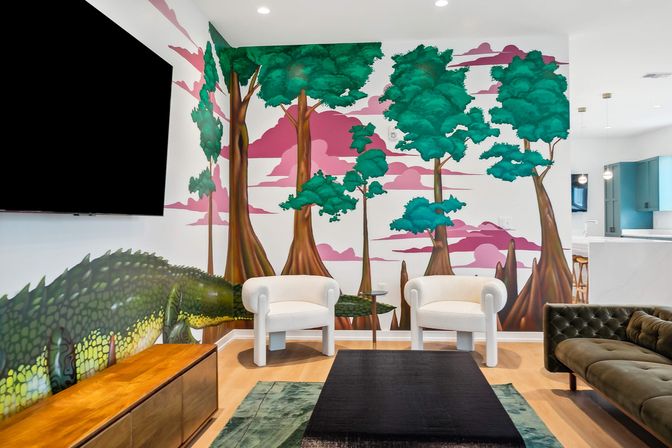 Modern living room with colorful wall mural of stylized trees, pink sky and a large green reptile, two white armchairs, olive tufted sofa, wall-mounted TV and wooden media console.