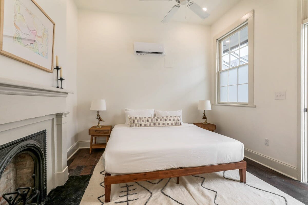 Bright, airy minimalist bedroom in a cozy urban home with a wooden platform bed, fireplace, hardwood floors, patterned rug, bedside tables with lamps, ceiling fan and wall-mounted AC.