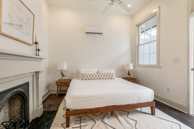 Bright, airy minimalist bedroom in a cozy urban home with a wooden platform bed, fireplace, hardwood floors, patterned rug, bedside tables with lamps, ceiling fan and wall-mounted AC.