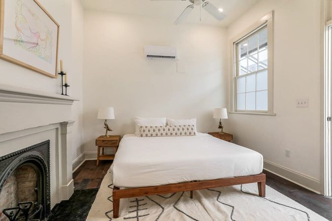 Bright, airy minimalist bedroom in a cozy urban home with a wooden platform bed, fireplace, hardwood floors, patterned rug, bedside tables with lamps, ceiling fan and wall-mounted AC.