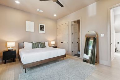 Contemporary master bedroom with king-size platform bed, white bedding and green accent pillows, black nightstands with lamps, arched full-length mirror, ceiling fan, light wood floors and neutral beige walls.