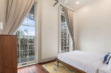 Sunlit minimalist bedroom with a low wooden bed dressed in white linens, tall French doors with beige curtains opening to a decorative wrought-iron balcony, dark hardwood floors and a yellow patterned rug.