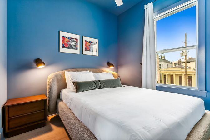 Cozy blue-walled bedroom with upholstered bed dressed in crisp white linens and a green bolster, wall-mounted reading lamps, wooden nightstand, pop-art prints above the headboard, and a large window framing a sunny city view of yellow neighboring buildings.