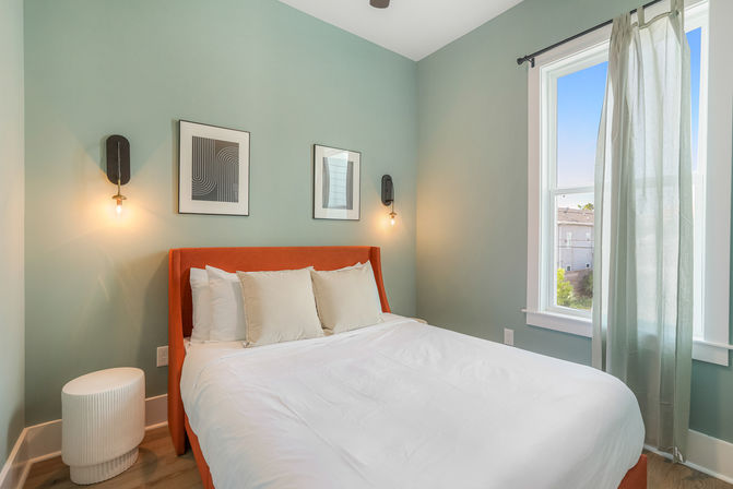 Light-filled modern apartment bedroom with teal walls, orange upholstered headboard, white bedding, two abstract framed prints, exposed-bulb wall sconces, sheer window curtain and round white nightstand